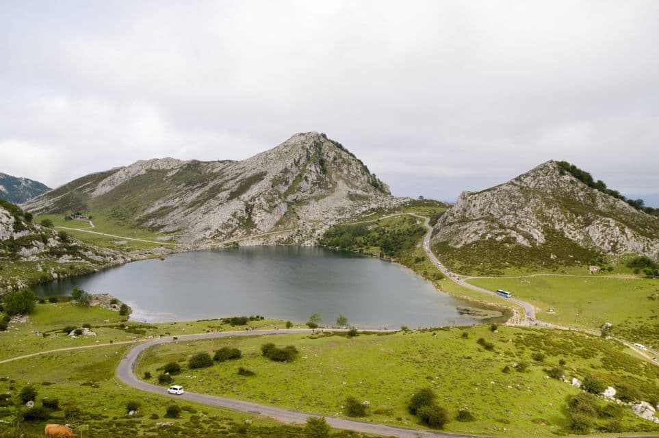 Una vista aérea de un lago rodeado de colinas verdes y montañas rocosas, con carreteras sinuosas y algunos vehículos bajo un cielo cubierto.