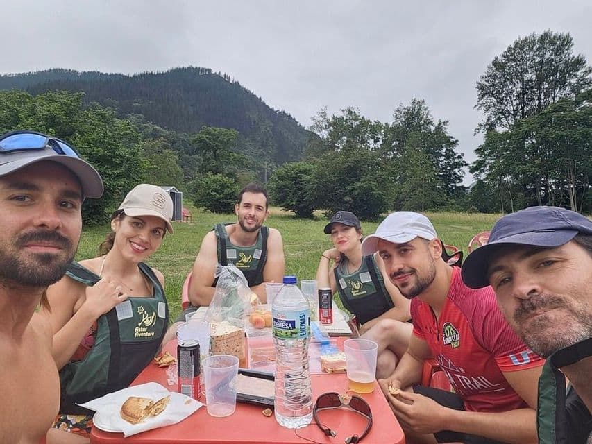 Un grupo de seis amigos de WeRoad, equipados con chalecos salvavidas, disfrutan de un pícnic en una mesa roja, ubicada en un campo de hierba con una colina boscosa de fondo.