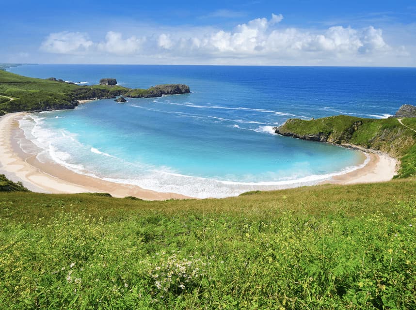 Vista aérea de una playa de arena con forma de media luna y una bahía turquesa, rodeada de acantilados verdes bajo un cielo azul.
