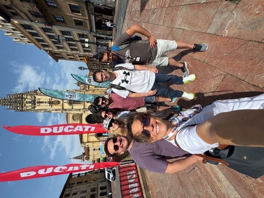 Un grupo de WeRoad se toma una selfie en una soleada plaza de la ciudad con una catedral y banderas rojas de Ducati de fondo.