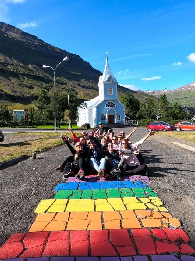 A WeRoad group trip poses for a photo on a rainbow-colored path in front of a light blue church at the base of a mountain.