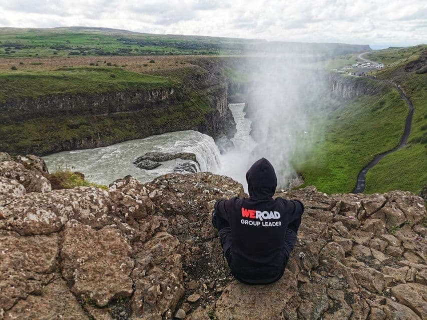 Ein WeRoad-Gruppenleiter in einem schwarzen Kapuzenpullover sitzt auf einer felsigen Klippe und blickt auf einen mächtigen Wasserfall in einer grünen Schlucht.