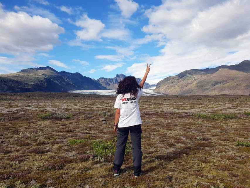 A WeRoad group leader from behind making a peace sign towards a glacier and mountains under a cloudy sky.