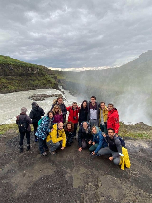 Un gruppo WeRoad in posa insieme su un dirupo accanto a una grande cascata nebbiosa sotto un cielo nuvoloso.
