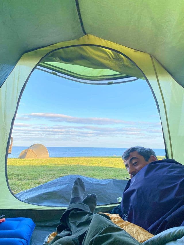 Un hombre sonriendo desde su saco de dormir dentro de una tienda de campaña, con vista al mar y otra tienda a través de la puerta abierta.