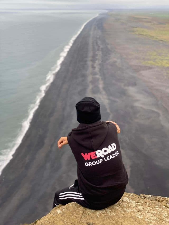 A WeRoad Group Leader sits on a rocky cliff overlooking a vast black sand beach stretching along the coastline.
