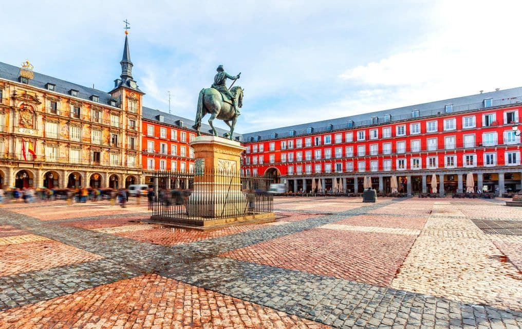 A bronze equestrian statue stands in the center of a large cobblestone plaza, surrounded by historic red buildings as people walk by.