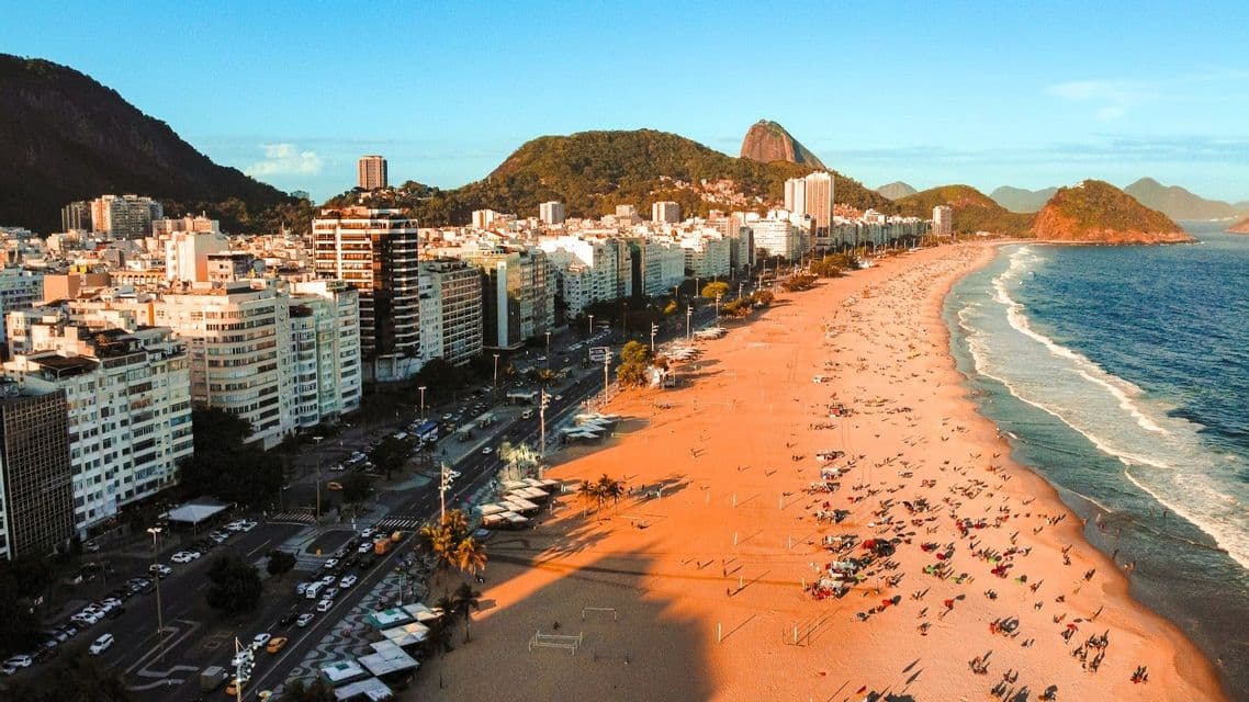 Eine Luftaufnahme eines belebten Stadtstrandes zur goldenen Stunde, mit hohen Gebäuden und grünen Bergen, die das Meer und den Sand säumen.