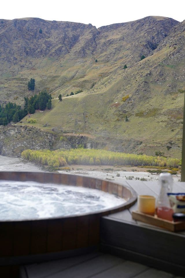 Una tinozza idromassaggio in legno con acqua gorgogliante su una terrazza, con vista su una valle fluviale e un fianco di montagna erboso.