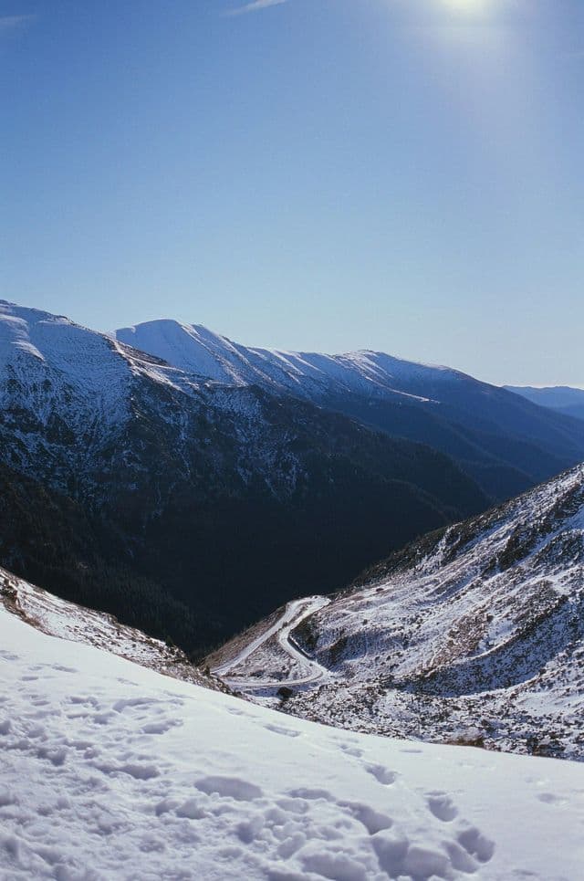 Una vista da un pendio innevato che domina una valle profonda dove una strada si snoda, con montagne innevate sullo sfondo sotto un cielo limpido.