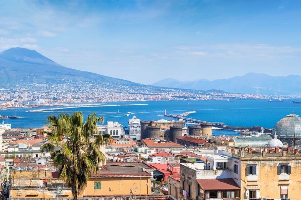 Ein Blick über die Dächer einer Küstenstadt, mit einer Bucht voller Boote und einem großen Vulkan, der sich in der Ferne unter blauem Himmel erhebt.