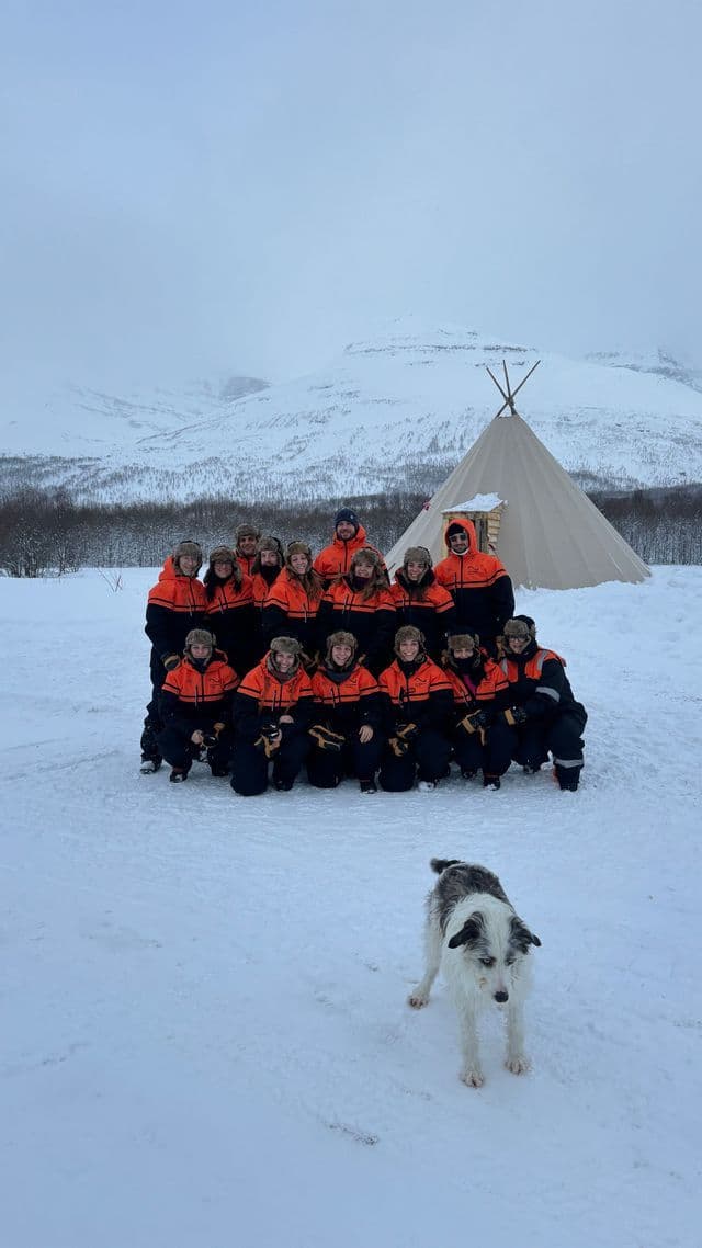 Un groupe WeRoad en voyage, vêtu d'équipements d'hiver assortis, prend la pose dans un paysage enneigé, avec un chien au premier plan, un tipi et des montagnes en arrière-plan.