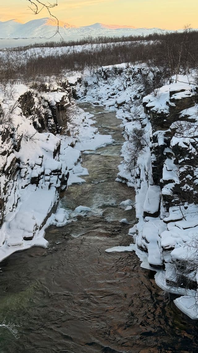 Un fiume scorre attraverso un canyon profondo e innevato con pareti rocciose, mentre montagne innevate sono visibili in lontananza.