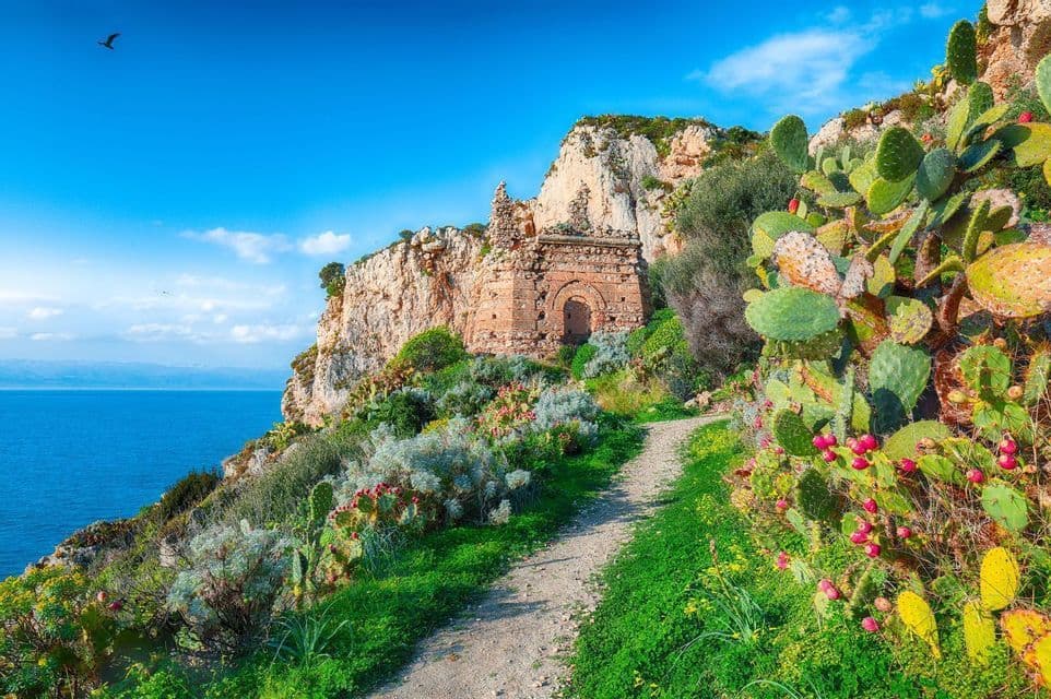 Un chemin de terre serpente le long de cactus figuiers de Barbarie sur une falaise, menant à une ruine en pierre surplombant la mer.