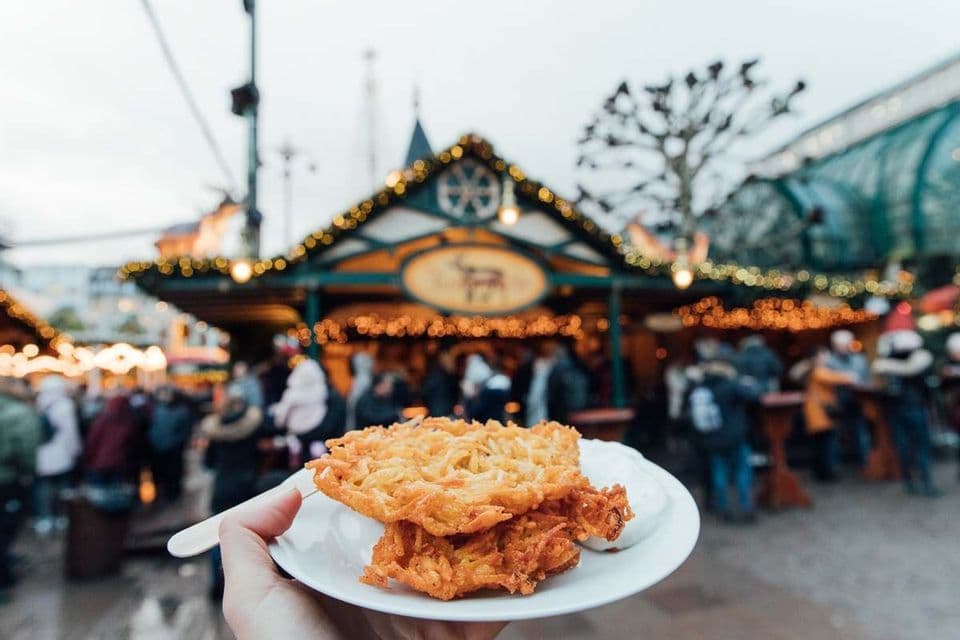 A hand holds a plate with fried potato pancakes at an outdoor Christmas market with decorated stalls and people in the background.