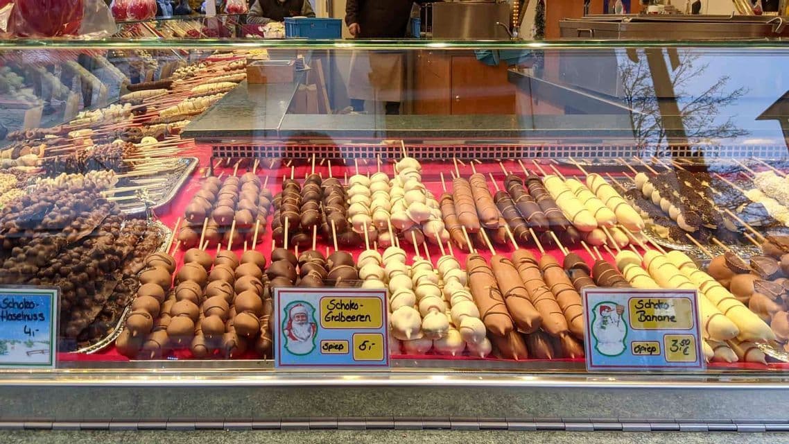 A variety of chocolate-covered fruits on skewers, including strawberries and bananas, are neatly arranged in a glass display case at a market stall.