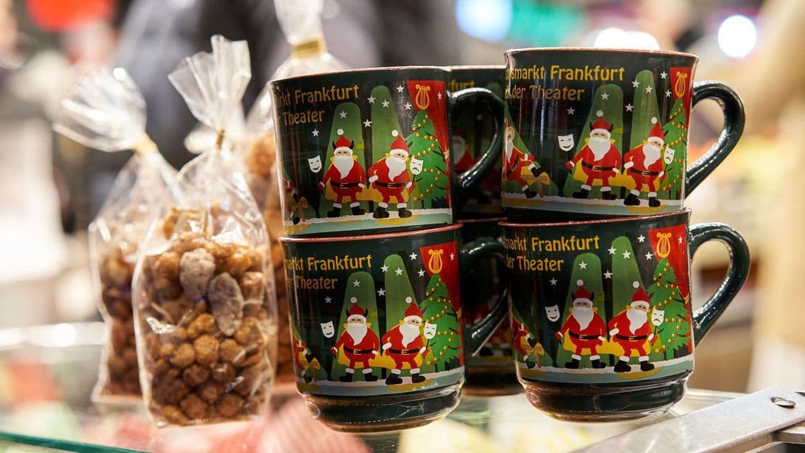 A close-up of stacked green Christmas-themed mugs with Santa Claus illustrations, next to bags of nuts on a shelf.