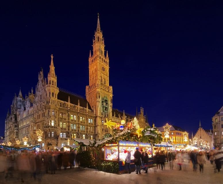 A bustling Christmas market with illuminated stalls in a snowy square at night, set against a large, lit-up Gothic building.
