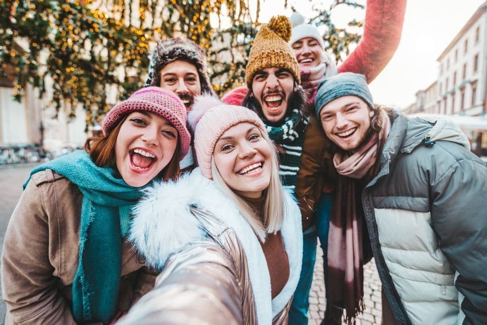 A WeRoad group trip of smiling friends in winter hats and jackets taking a selfie on a city street with festive lights.