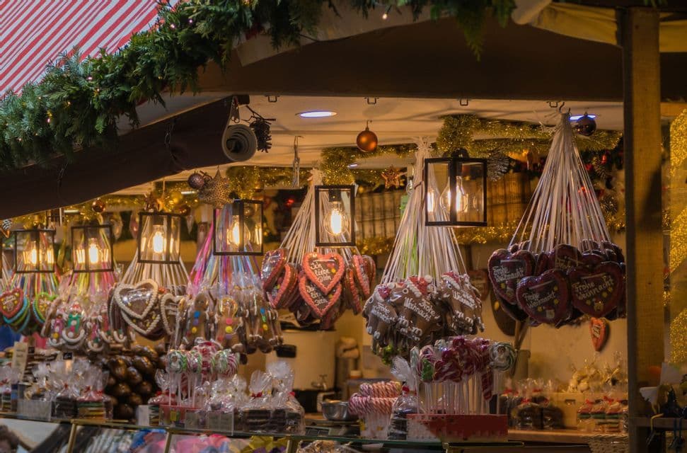 A Christmas market stall displays gingerbread hearts, candies, and lollipops under warm lanterns and festive garlands.