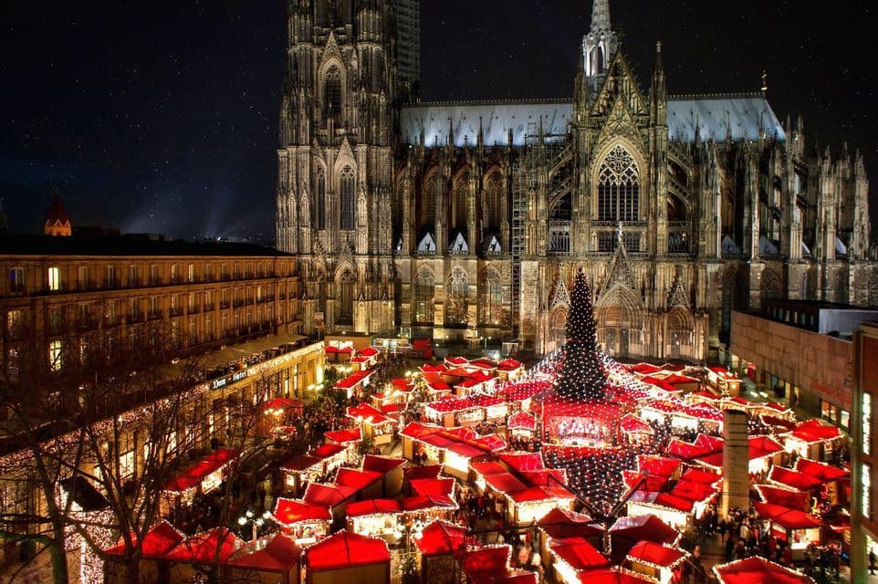 A Christmas market with glowing red stalls and a large tree bustling with people in front of a Gothic cathedral at night.