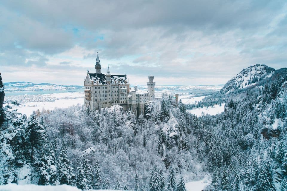 A white castle on a hill is surrounded by a vast forest and mountains, all covered in snow under a cloudy sky.