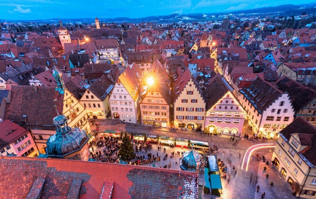 An aerial view of a crowded Christmas market in a historic town square at dusk, with lights illuminating the traditional buildings.