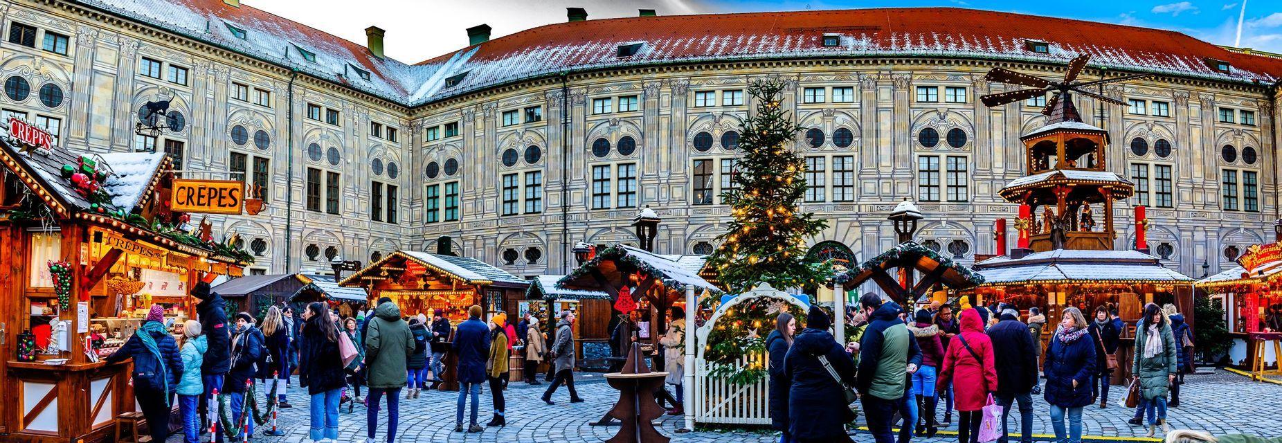 A busy Christmas market with festive stalls and a large tree in a cobblestone square, in front of a grand, ornate building.