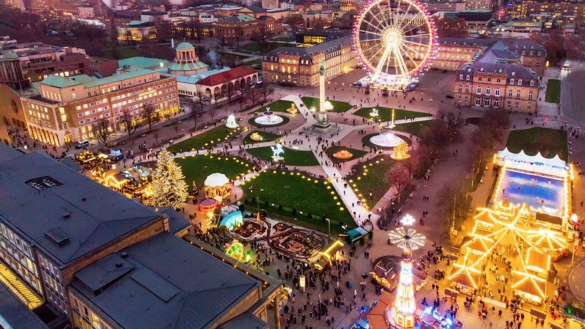 An aerial view of a large city square illuminated for a Christmas market, with a Ferris wheel, an ice rink, and crowds of people.