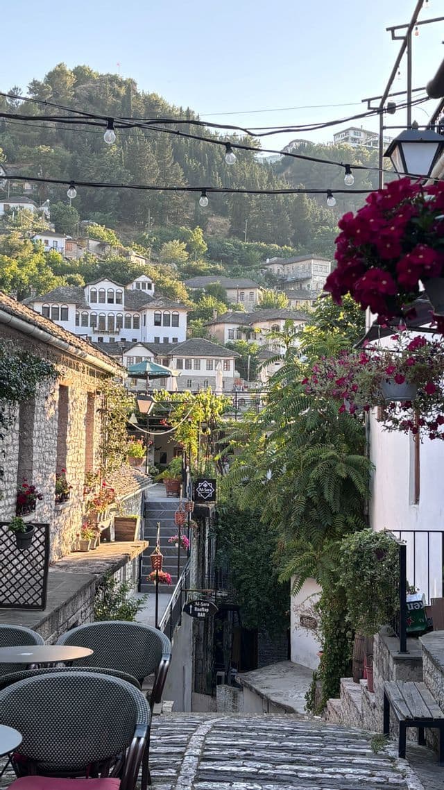 Une vue sur une étroite ruelle pavée dans une ville européenne à flanc de colline avec des bâtiments en pierre, une végétation luxuriante et des guirlandes lumineuses.