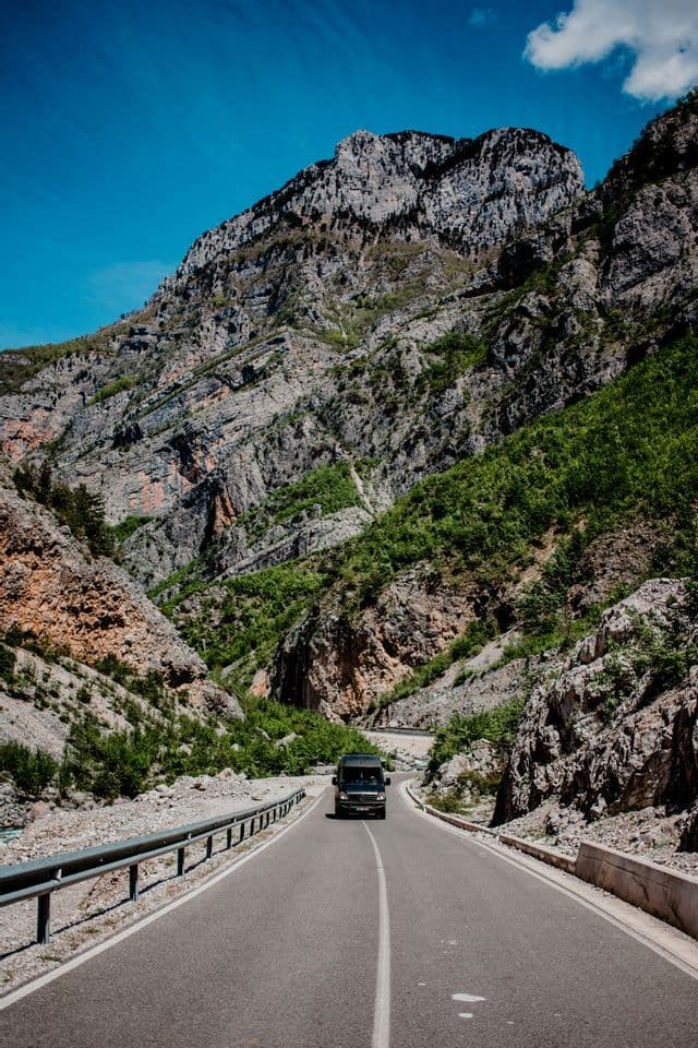 Un van sombre roule sur une route sinueuse à travers une vallée montagneuse rocheuse sous un ciel bleu clair.