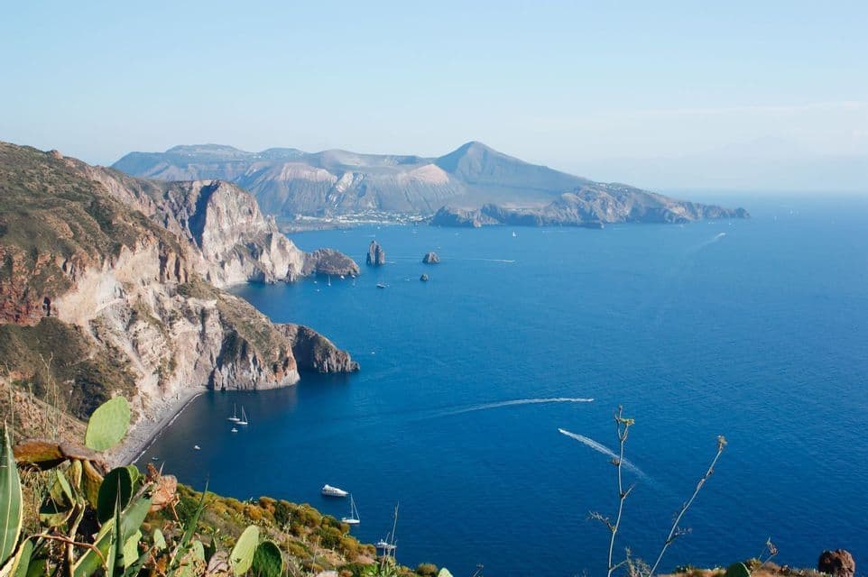 Vue plongeante d'un littoral rocheux et volcanique longeant la mer d'un bleu profond, où plusieurs bateaux naviguent près d'îles lointaines sous un ciel clair.