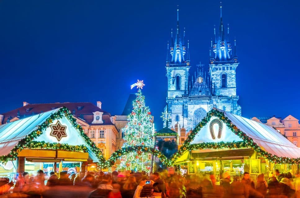 A bustling Christmas market at night, featuring an illuminated tree, festive stalls, and a large Gothic church in the background.