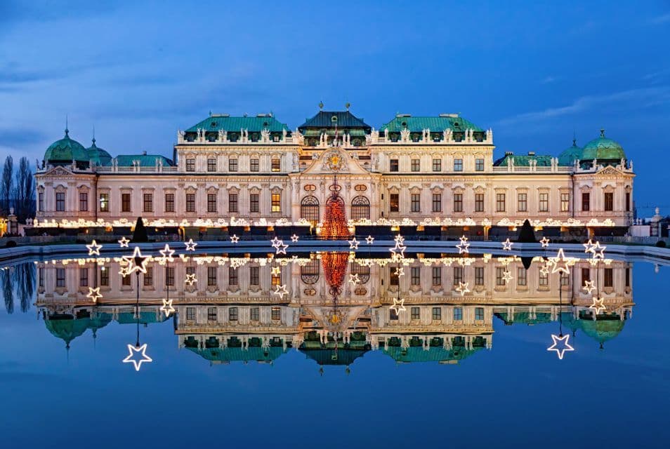 An illuminated palace with Christmas decorations, including glowing stars, is reflected in a still pool at dusk.