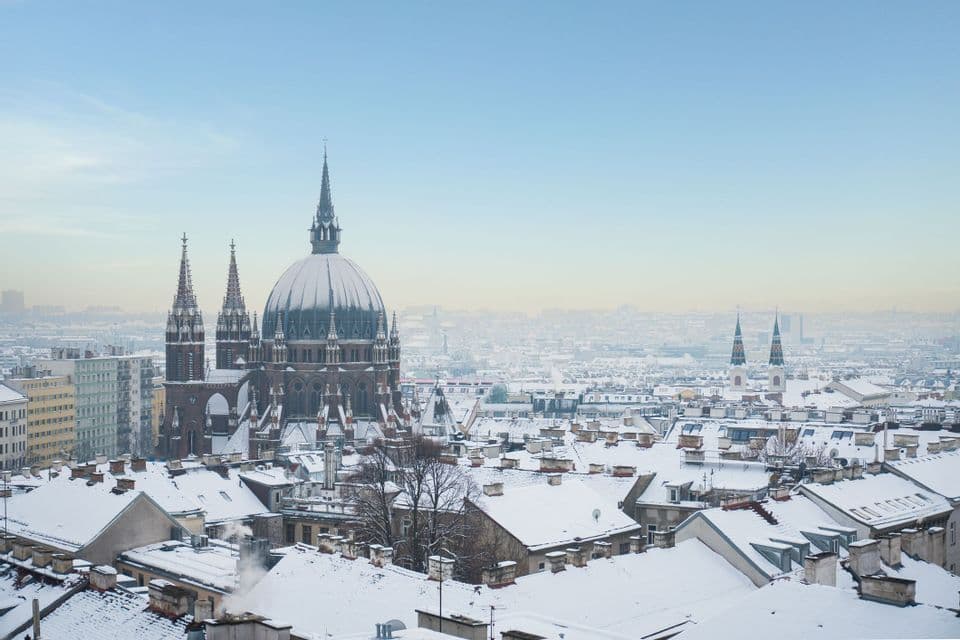A large, domed cathedral overlooks a cityscape of snow-covered rooftops under a pale blue sky.