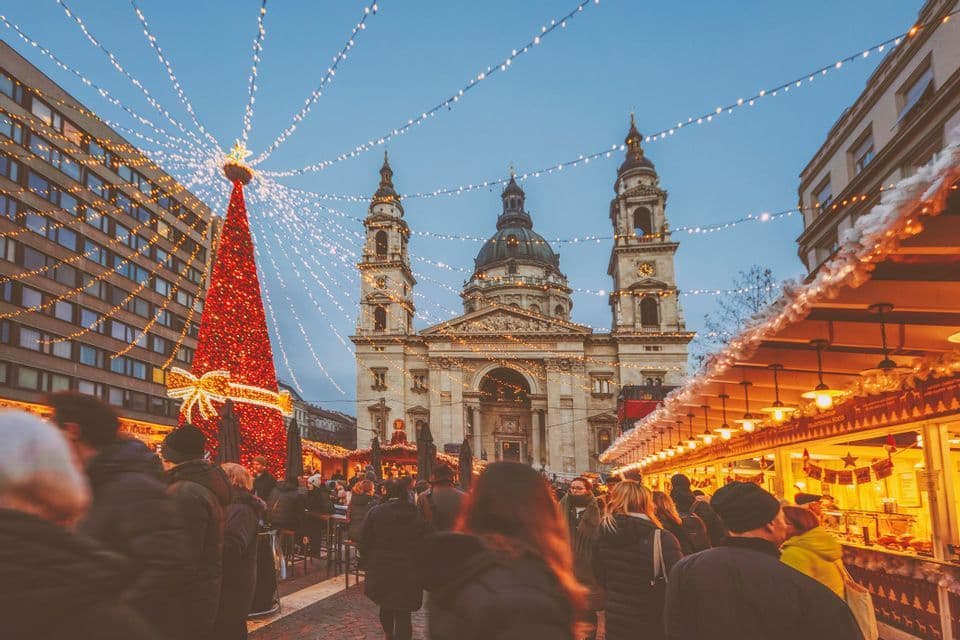 A crowd of people at a Christmas market at dusk, with an illuminated tree and an ornate basilica under strings of lights.
