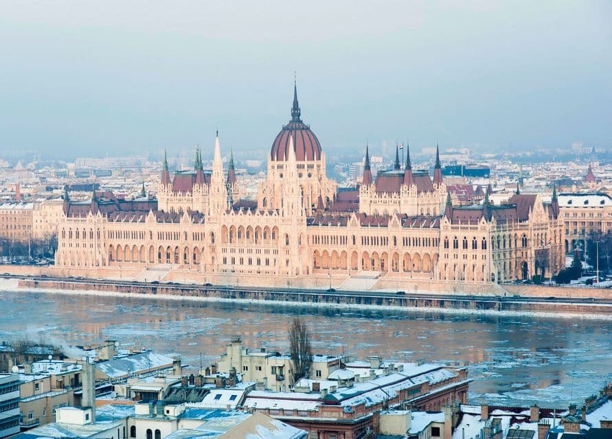 A grand, gothic-style building with a central dome and spires sits along an icy river in a snow-dusted city.