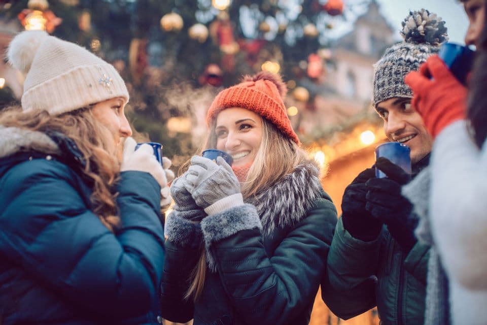 A WeRoad group trip of friends in winter hats and coats, smiling and drinking hot beverages from mugs at an outdoor market.