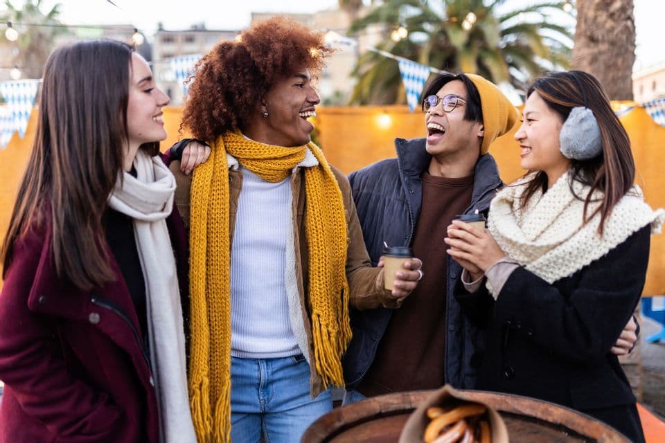 A WeRoad group trip of four friends laughing together and holding coffee cups at an outdoor winter market.