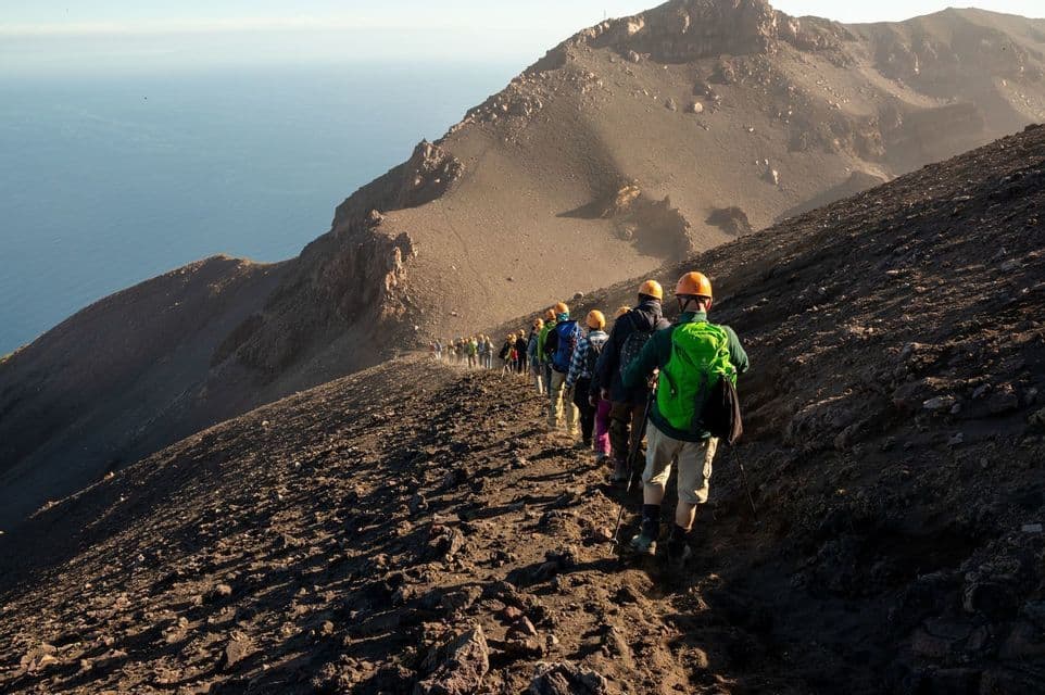 Un groupe WeRoad, casqué, randonne en file indienne sur un sentier volcanique accidenté, surplombant la mer.