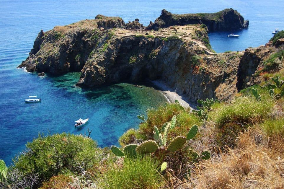 Eine Draufsicht auf eine felsige Bucht mit einem kleinen Kiesstrand, klarem türkisfarbenem Wasser und mehreren kleinen Booten, die vor Anker liegen.