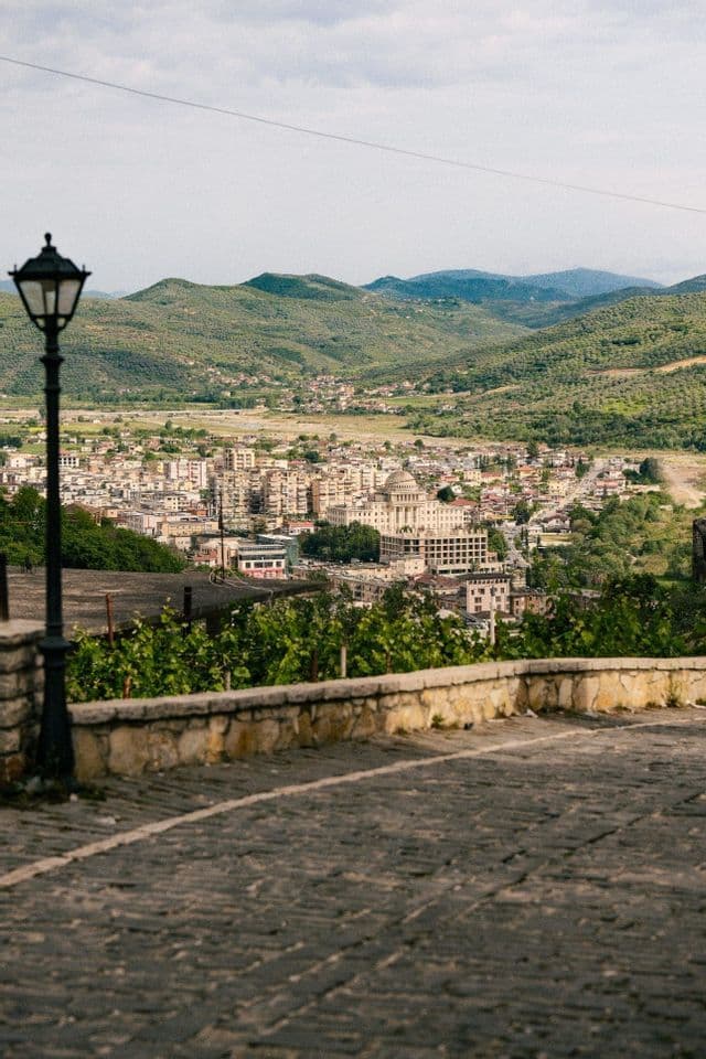 Una ciudad enclavada en un valle verde, vista desde un camino empedrado alto con una farola negra en primer plano.