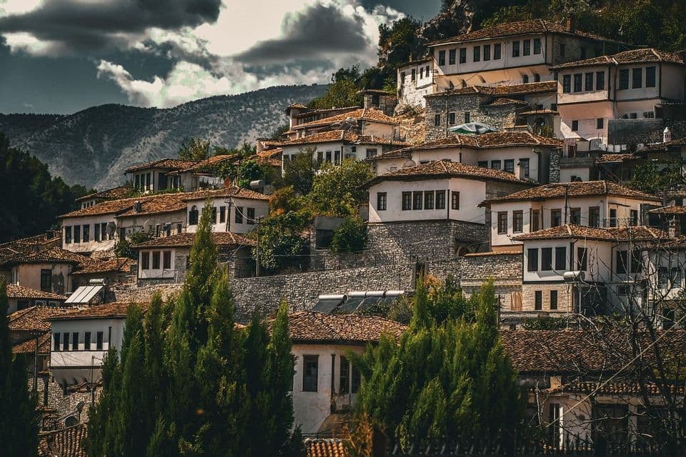 A village of traditional stone and white-walled houses with terracotta roofs built on a steep, tree-covered hillside under a cloudy sky.