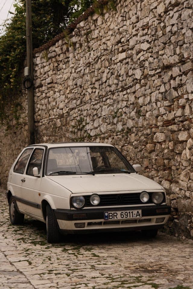 A white vintage car parked on a cobblestone street next to a high, rustic stone wall.