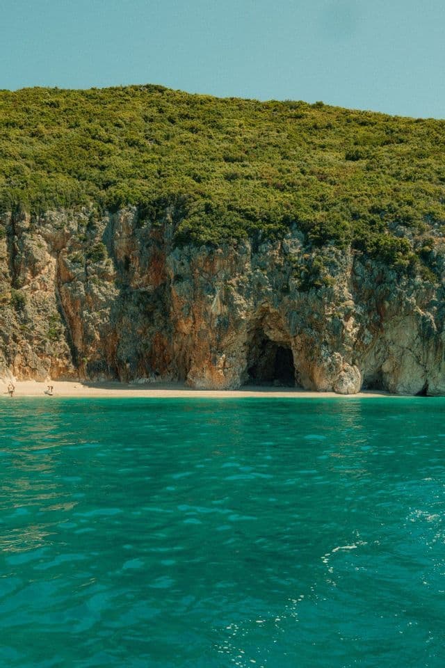 A view from the turquoise sea of a secluded sandy beach with a cave at the base of a lush, rocky cliff.