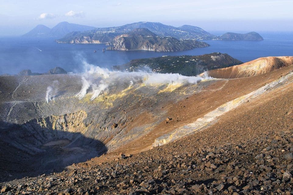 Une vue plongeante depuis un cratère volcanique avec de la vapeur s'échappant des fumerolles, surplombant une vaste mer avec plusieurs îles au loin.
