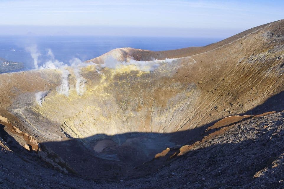 Vue large d'un cratère volcanique avec des dépôts de soufre jaune et de la vapeur s'échappant des évents. La mer est visible au loin.