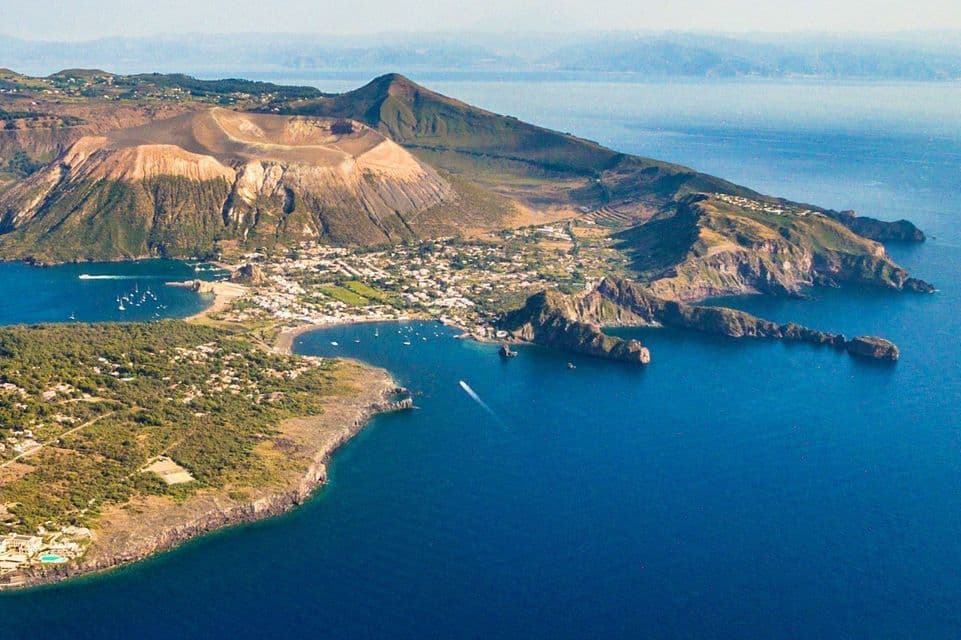 Veduta aerea di un'isola vulcanica caratterizzata da un ampio cratere, con un paese e imbarcazioni in una baia lungo la costa.