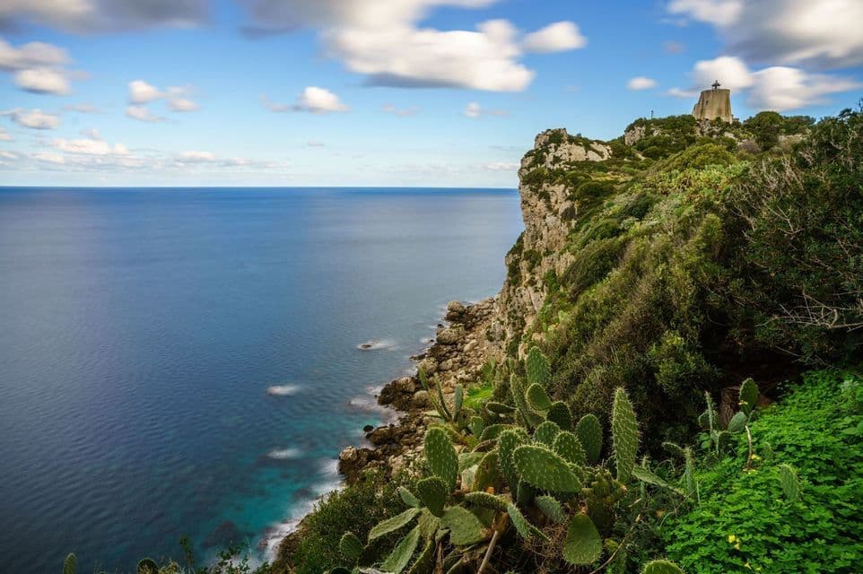 Una torre di pietra si erge su una scogliera rocciosa e rigogliosa, affacciata sull'oceano blu intenso sotto un cielo parzialmente nuvoloso.