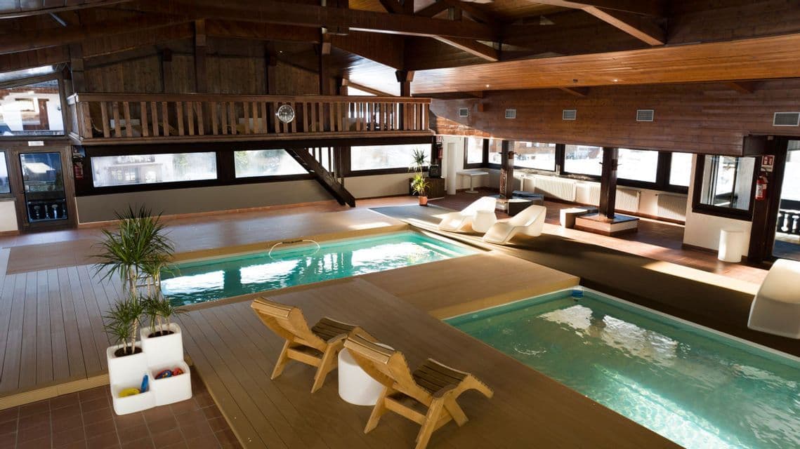 Un'area benessere interna con due piscine, pavimentazione in legno e lettini sotto un alto soffitto con travi a vista.